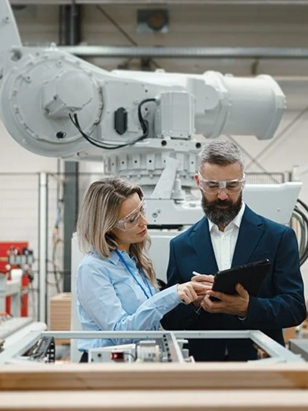 Deux personnes discutent et consultent une tablette dans une usine automatisée, devant un grand bras robotisé.