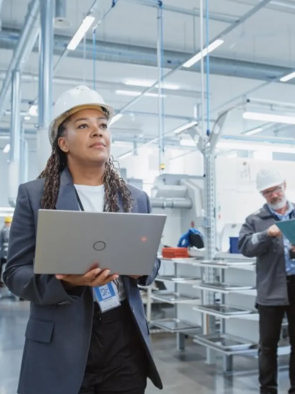 Personne portant un casque de sécurité utilisant un ordinateur portable dans une usine moderne, avec d’autres travailleurs examinant des documents en arrière‑plan. 