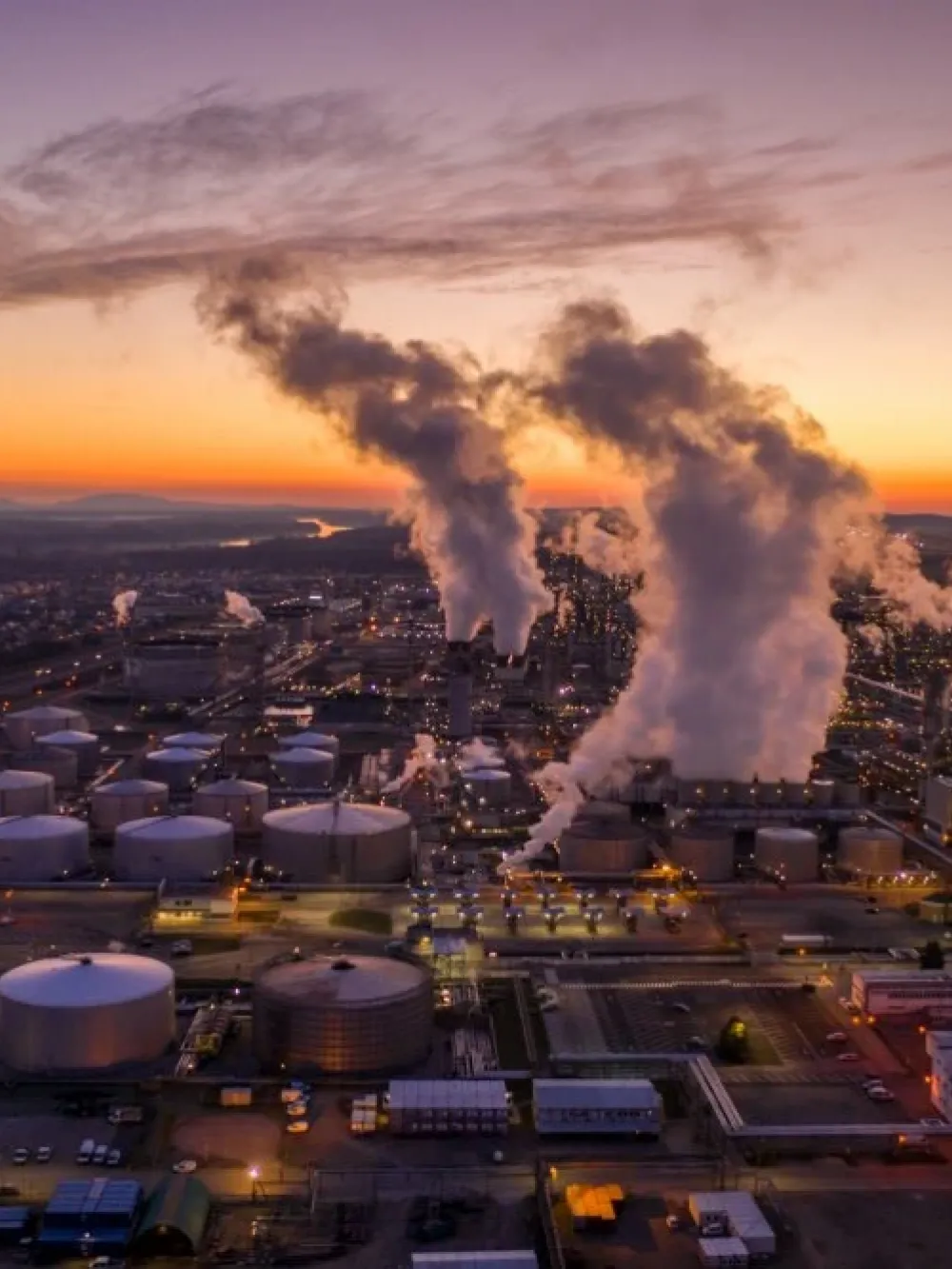 Vue aérienne d’un vaste complexe industriel avec des réservoirs de stockage et plusieurs cheminées dégageant de la vapeur au lever du soleil. 