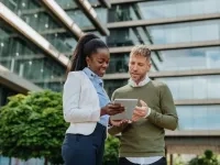 Deux personnes discutent ensemble à l’extérieur d’un immeuble moderne en consultant une tablette. 