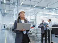 Personne portant un casque de sécurité utilisant un ordinateur portable dans une usine moderne, avec d’autres travailleurs examinant des documents en arrière‑plan. 