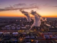 Vue aérienne d’un vaste complexe industriel avec des réservoirs de stockage et plusieurs cheminées dégageant de la vapeur au lever du soleil. 